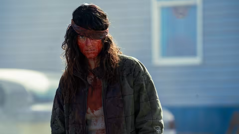 a bloodied person with long hair and headband standing outside