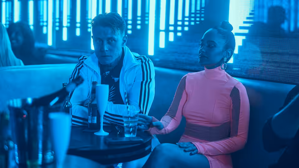 Preston (George MacKay) and a woman in pink seated in a blue-lit bar