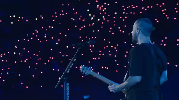 A bald guitarist plays onstage before a crowd dotted with glowing lights