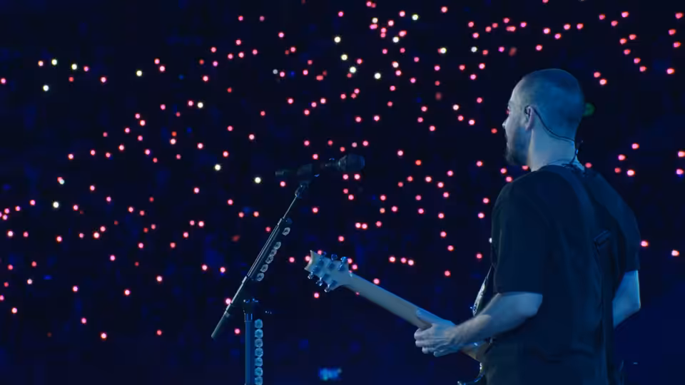 A bald guitarist plays onstage before a crowd dotted with glowing lights
