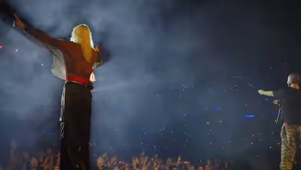 A blonde performer raises an arm toward the crowd as a guitarist plays through stage smoke
