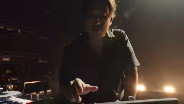 A musician leans over a keyboard or mixing console under warm stage lights