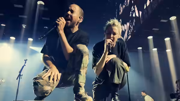 A male singer and a blonde singer crouch at the stage edge, singing into microphones
