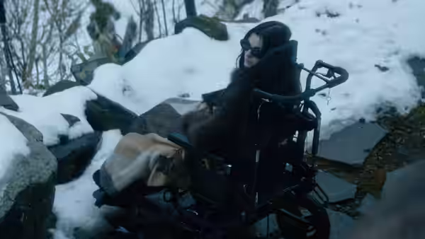 a woman in sunglasses and a fur coat sitting in a wheelchair on snowy rocks