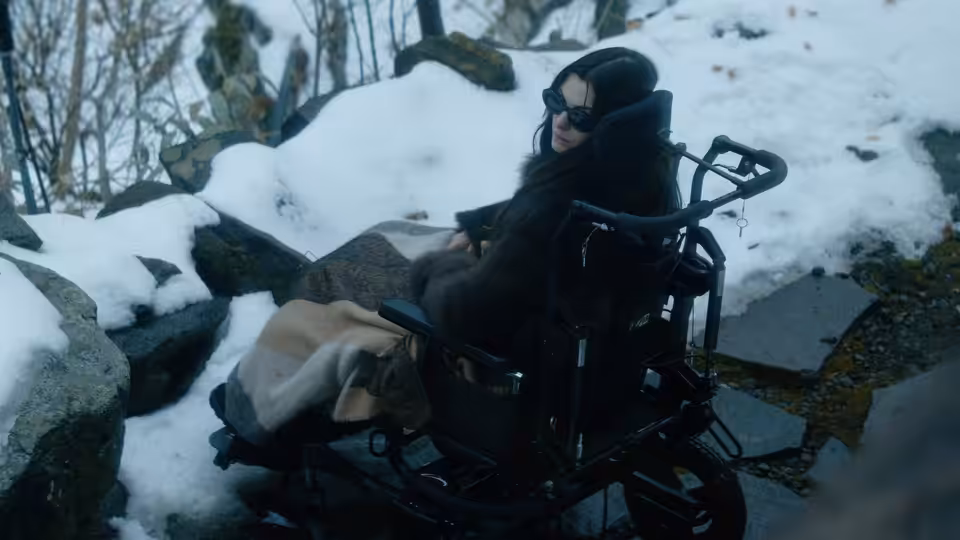 a woman in sunglasses and a fur coat sitting in a wheelchair on snowy rocks