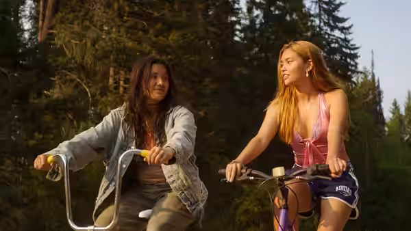 Two women ride bicycles side by side along a path through a forest