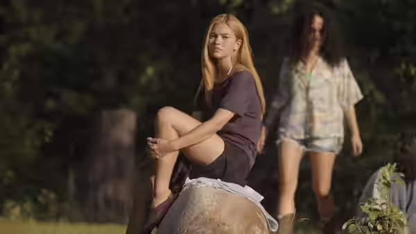 A woman with long red hair sits on a rock while another stands in the background