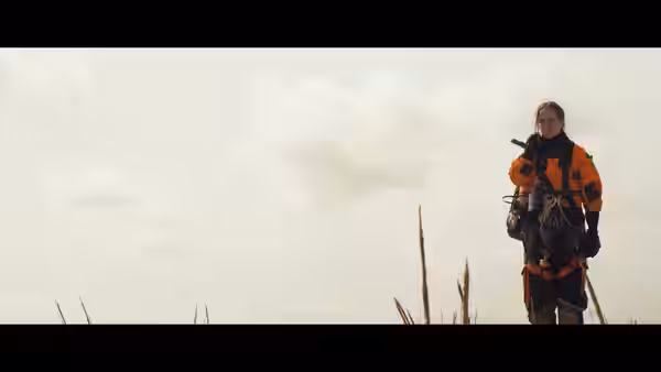 A person in orange outdoor gear stands in tall grass, holding a handheld radio