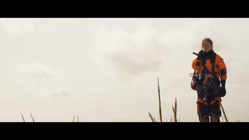 A person in orange outdoor gear stands in tall grass, holding a handheld radio