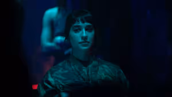 A woman in a camouflage jacket stands beneath blue lighting, with another figure behind her