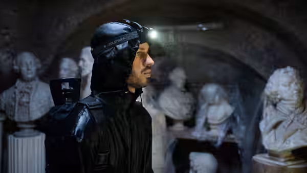 A black-clad man wearing a headlamp stands among busts in a stone cellar