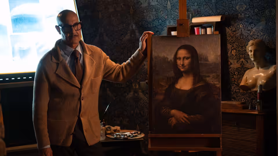 Roger (Stanley Tucci) stands beside an easel displaying the Mona Lisa in a furnished room