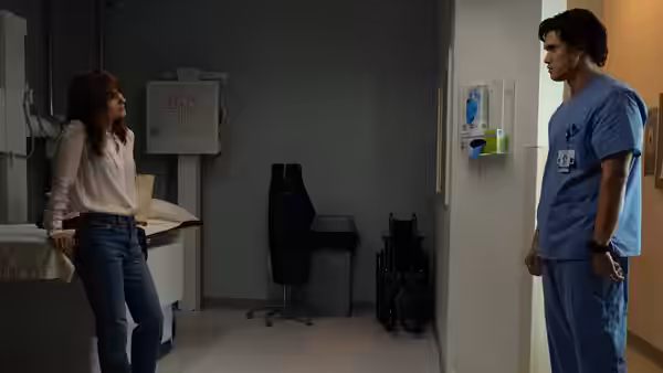 A woman leans against a table while speaking to a man in medical scrubs