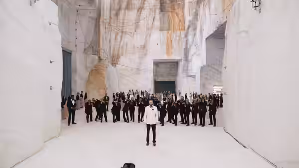 A figure in white addresses a large crowd inside a vast marble chamber
