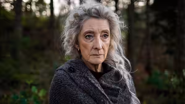 An older woman in a dark shawl stands among trees in a forest