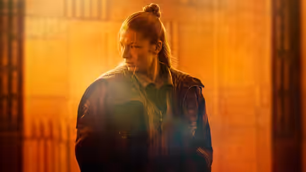 Cora (Hunter Schafer) standing alone in a warmly lit industrial space