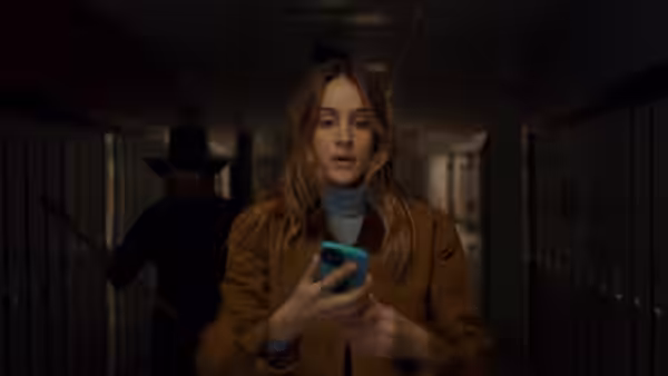 A young woman checks her phone in a school hallway as a figure approaches