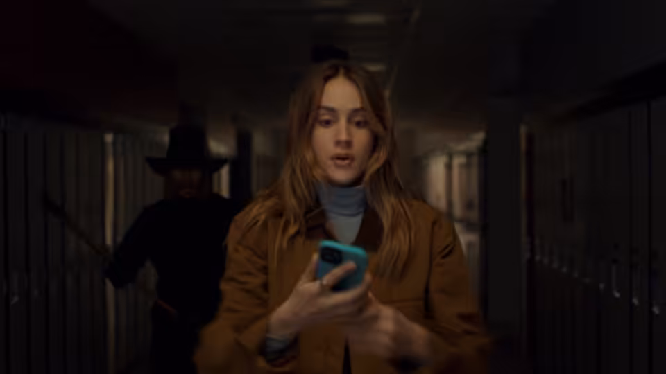 A young woman checks her phone in a school hallway as a figure approaches