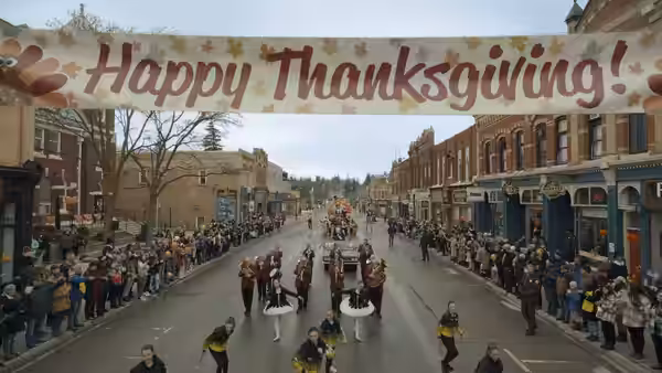 A parade marches down a street beneath a large Happy Thanksgiving banner