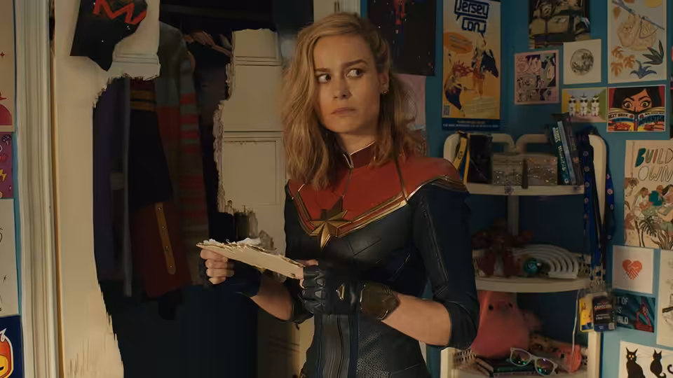 Carol Danvers (Brie Larson) holding a device in a poster-filled bedroom