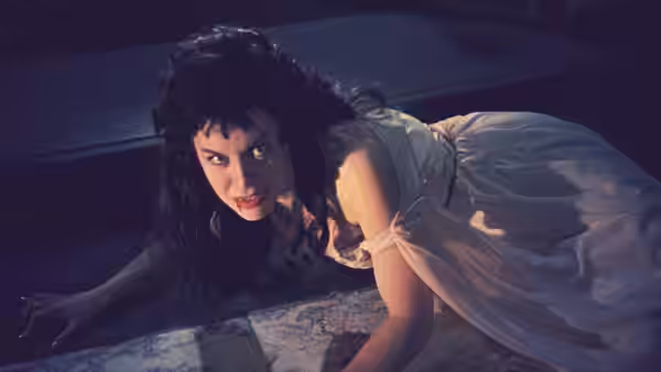 A vampire woman crouches over another woman lying on a bed