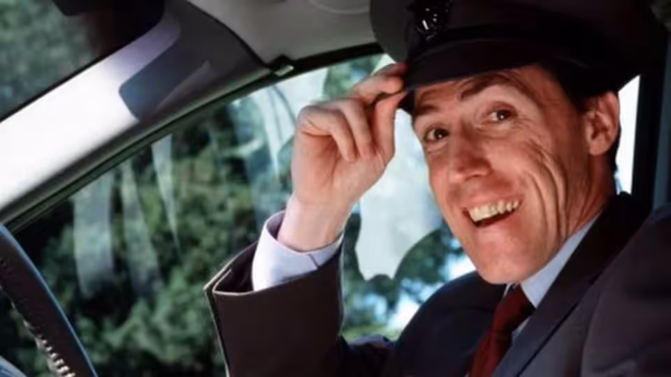 Keith (Rob Brydon) smiles and tips his cap from the driver's seat of a car