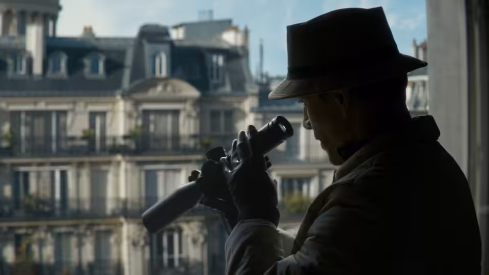 The Killer (Michael Fassbender) peering through binoculars from a window overlooking buildings