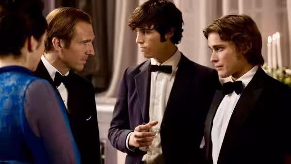 Three men in tuxedos converse closely at a formal indoor event with candles behind