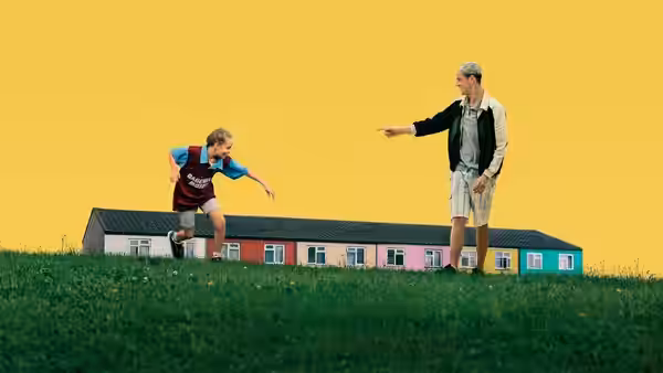 Georgie (Lola Campbell) running toward Jason (Harris Dickinson) on grass before colorful houses
