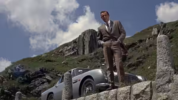 James Bond (Sean Connery) stands beside his Aston Martin on a rocky hillside