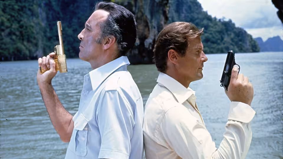 Scaramanga and Bond stand back to back holding guns by the water