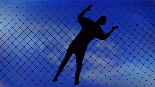 A silhouetted person reaches toward a chain-link net against a blue sky