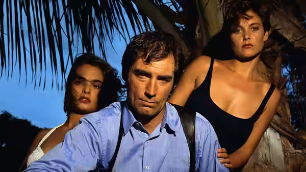 James Bond (Timothy Dalton) standing between two women under palm trees