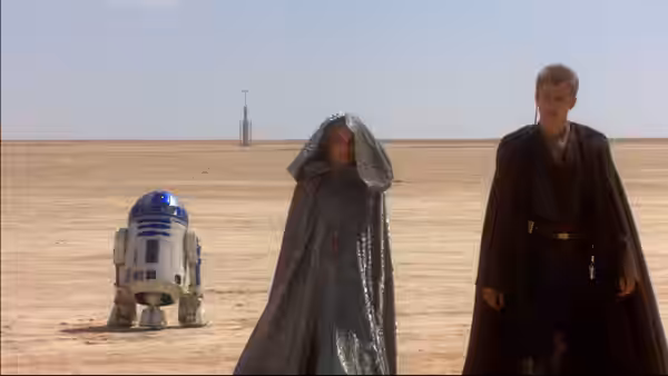 A cloaked woman a blue astromech droid and a robed man stand in the desert