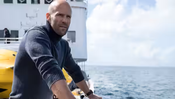 Jason Statham leans on the railing of a research vessel at sea