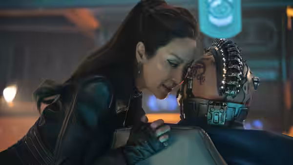 a woman in leather jacket leaning close to a cyborg helmeted figure