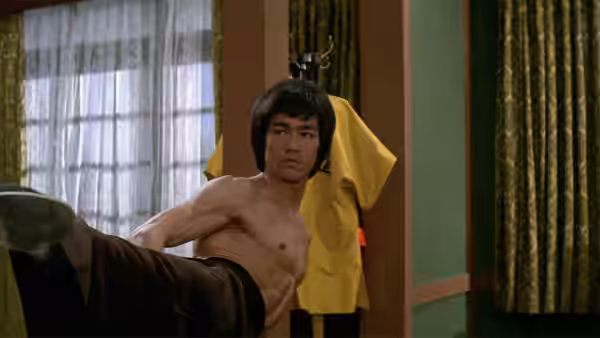Lee (Bruce Lee) extends a high kick inside a room with a yellow jacket hanging behind him