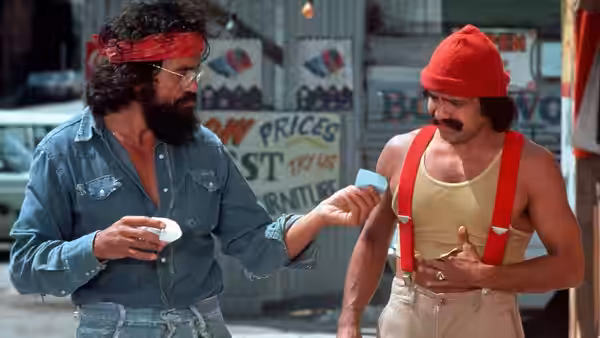 Man (Tommy Chong) offers Pedro (Cheech Marin) a small paper at an outdoor market