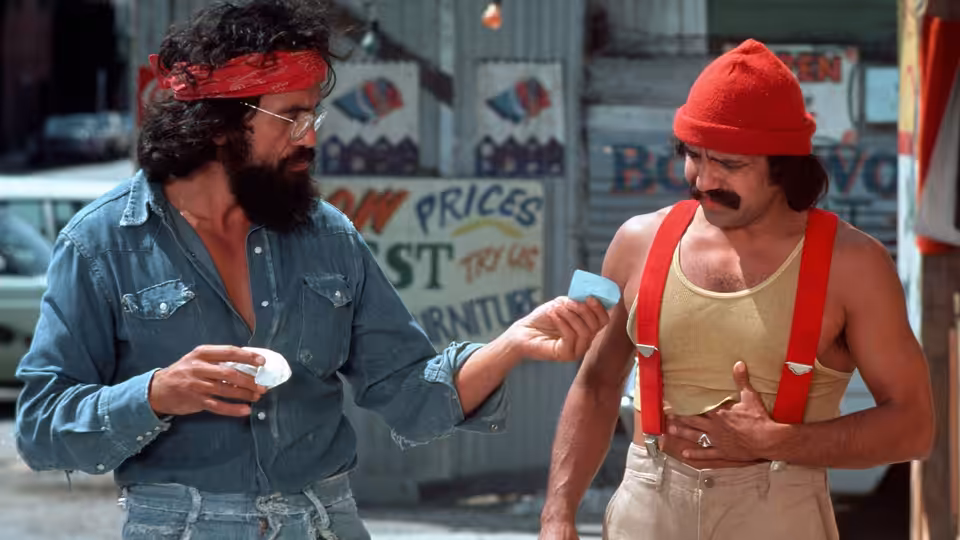 Man (Tommy Chong) offers Pedro (Cheech Marin) a small paper at an outdoor market