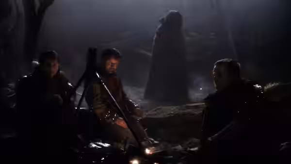 Three soldiers sit by a campfire as a cloaked figure stands behind them