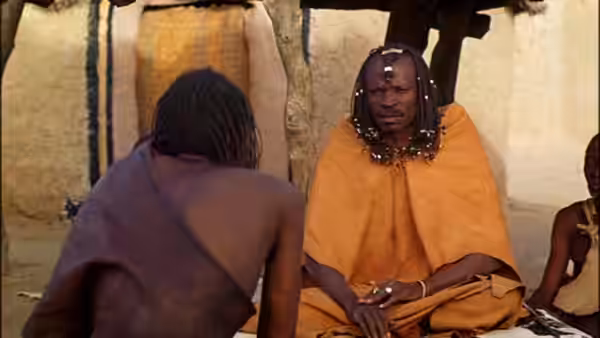 A seated man in an orange robe faces a bare-backed visitor inside a mud dwelling