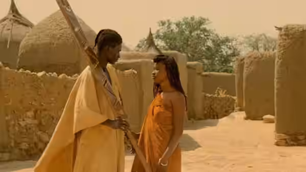 A man carrying a long staff speaks with a woman in an orange wrap
