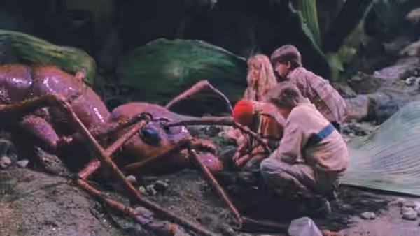Four tiny children crouch beside a giant ant among garden debris