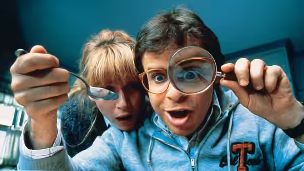 A boy peers through a magnifying glass while a girl watches indoors