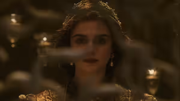 Isabel (Rachel Weisz) wears a jeweled crown and gold gown behind a glass pane