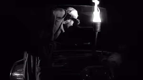 A hooded figure bends over a vehicle, holding a torch in the darkness