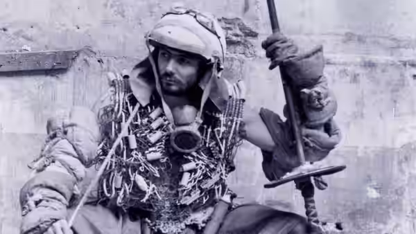 A helmeted scavenger covered in straps and gear grips a staff beside a ruined wall
