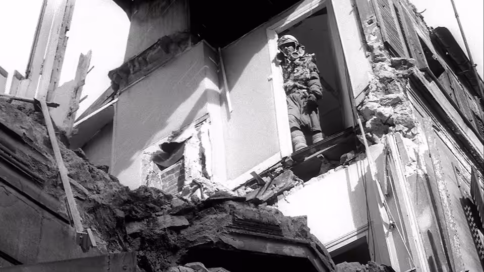 An armed man stands in a doorway amid the broken walls of a ruined building