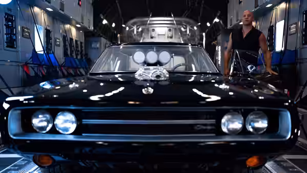 Dominic Toretto (Vin Diesel) stands beside a black muscle car in a large hangar
