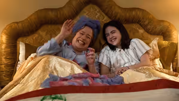Two girls in pajamas lie in bed smiling and waving